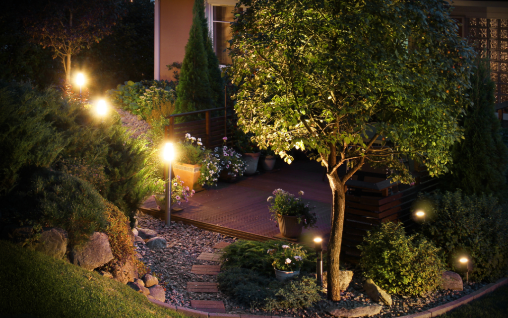 Landscape lighting throughout raised landscape bed with a tree, decorative stones, and walkway.